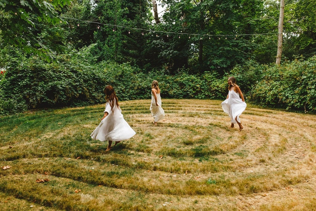 Three women in white dresses dancing barefoot on a grass labyrinth