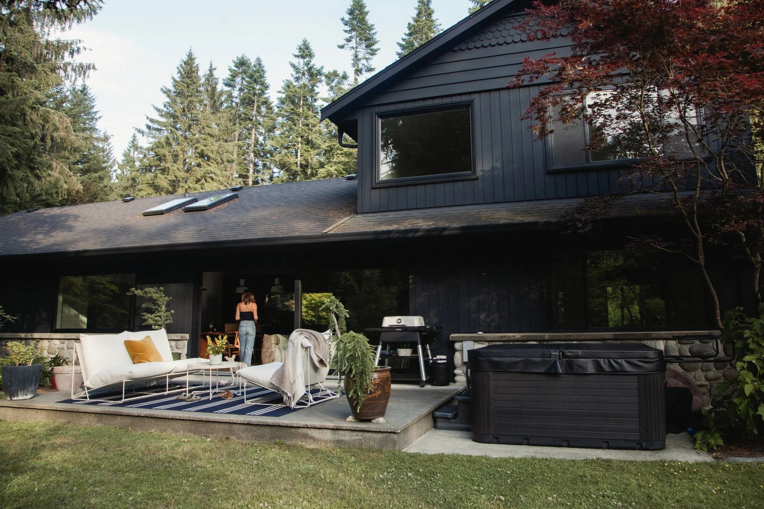 The black-sided house with a sofa and rugs on the deck below the trees