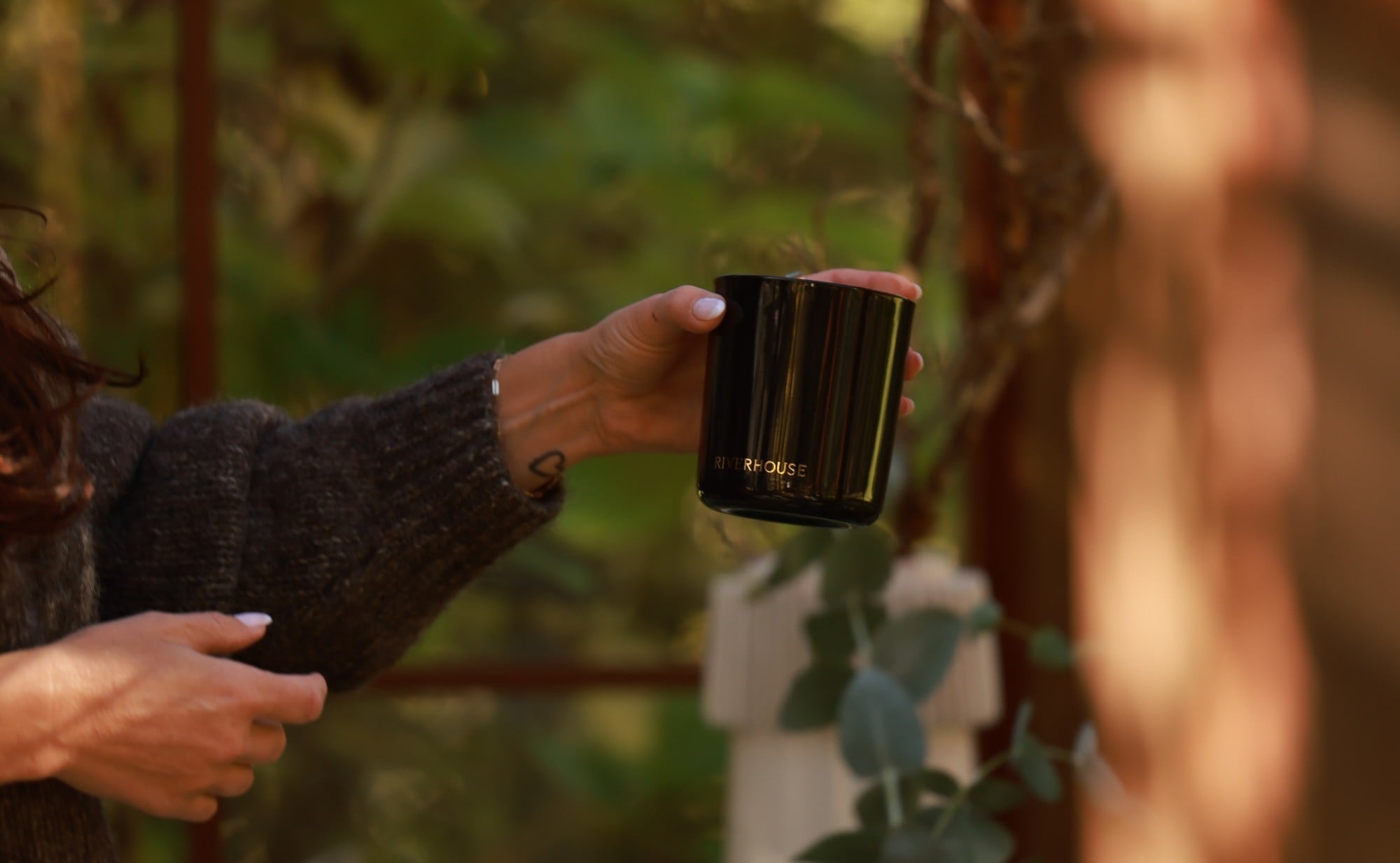 A hand holding a Riverhouse candle in the greenhouse